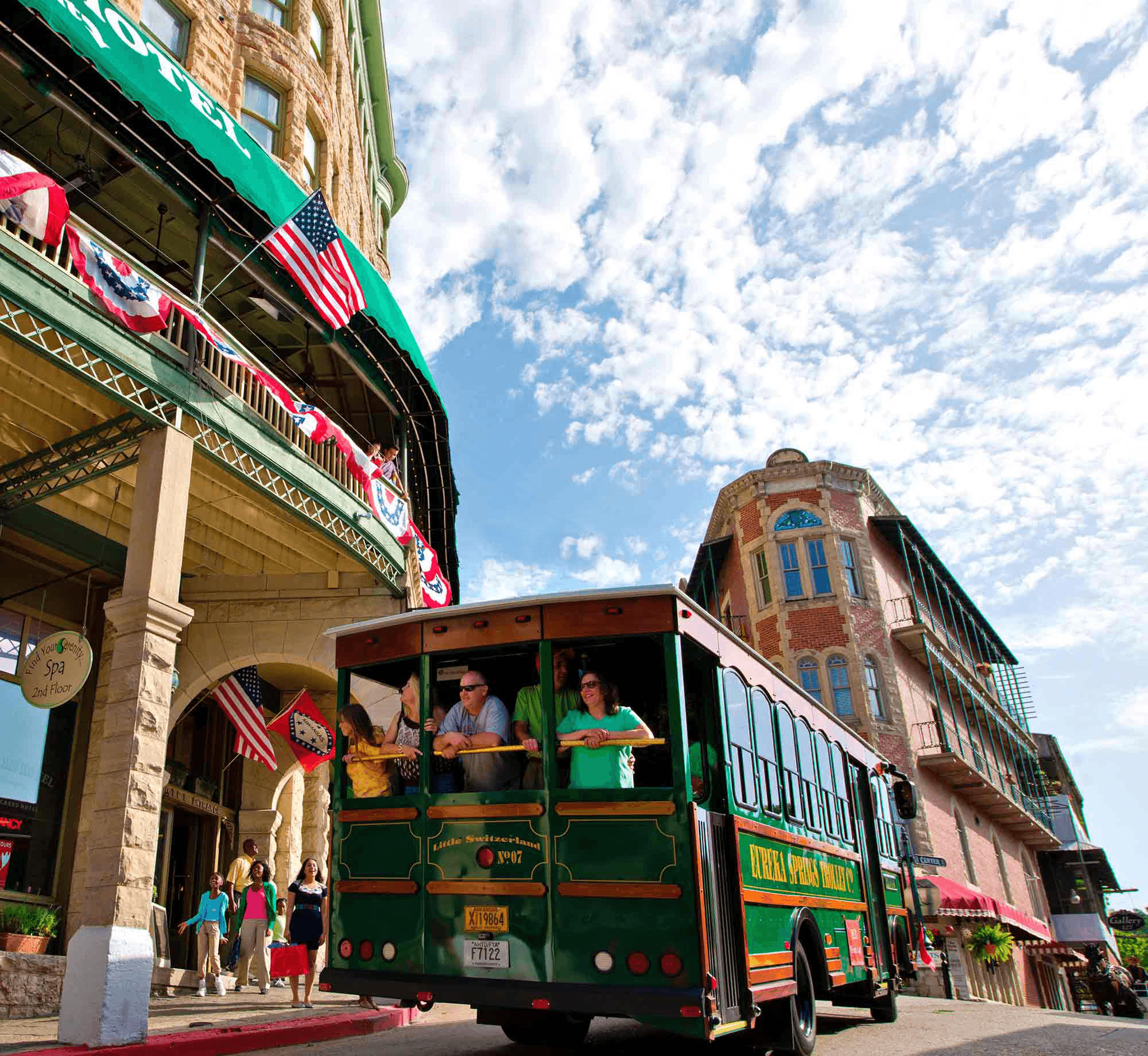 A historic green trolley with passengers passes the Basin Park Hotel and a red-brick building under a bright blue sky. American flags adorn the hotel.