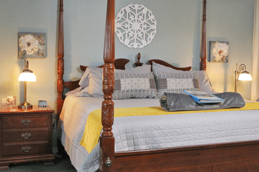 A cozy bedroom featuring a dark wooden four-poster bed with gray and yellow bedding. Two floral artworks hang above side tables with lamps.
