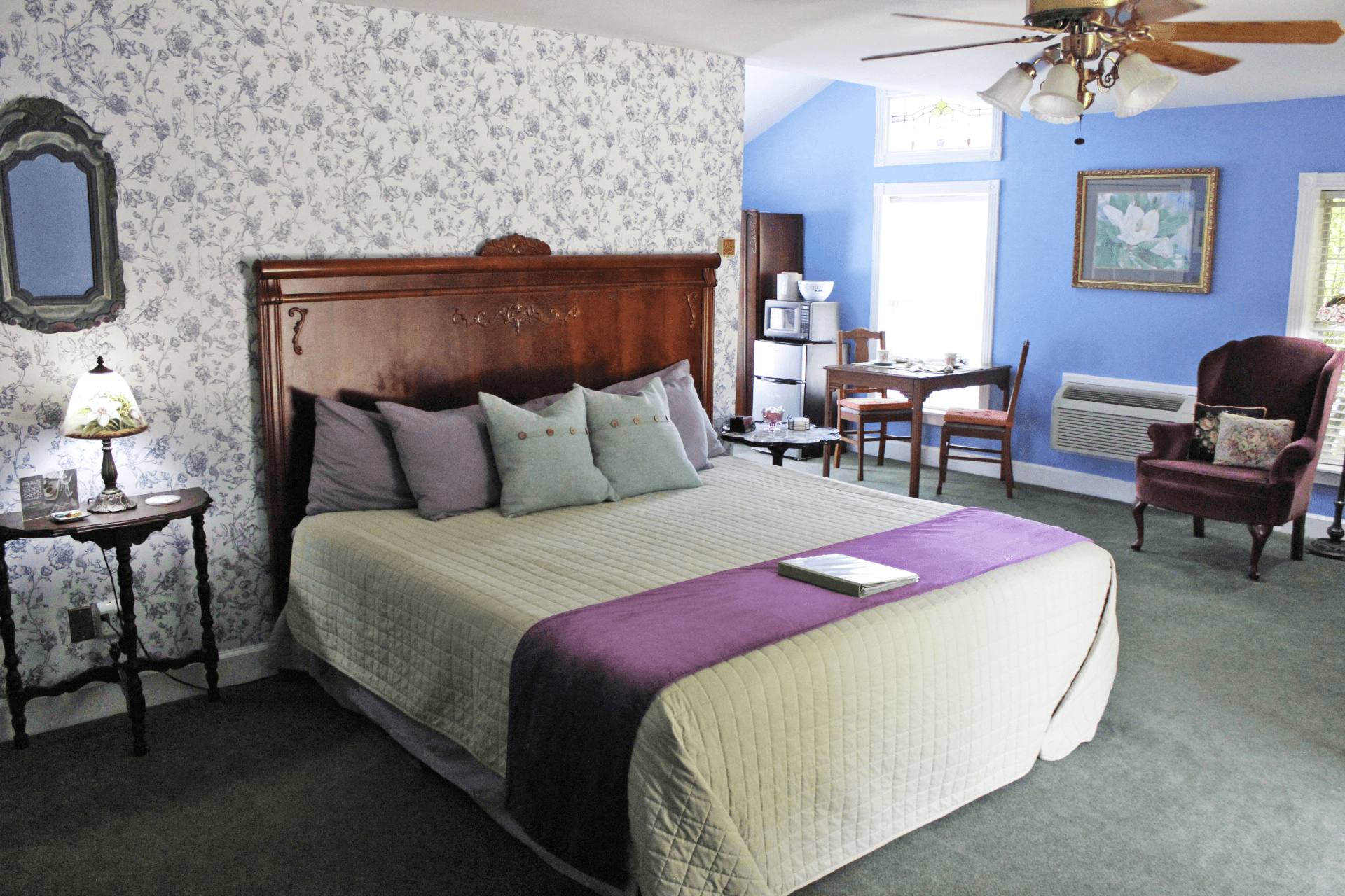Cozy bedroom with floral wallpaper and large bed featuring purple accents. Includes a vintage side table lamp, armchair, dining area, and ceiling fan.