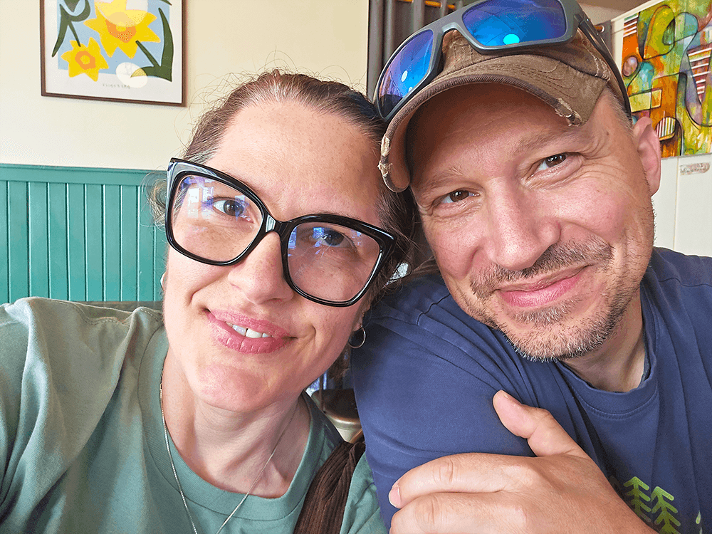 A close-up selfie of a woman with glasses and a man smiling in a brightly decorated café.